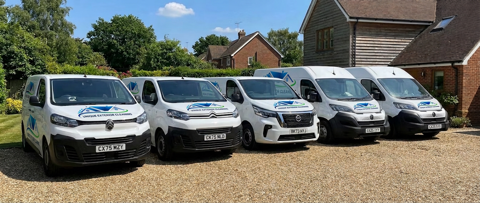 Five white Unique Exterior Cleaning vans parked in a row on a gravel driveway